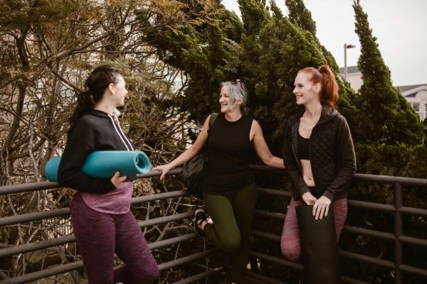 Three women with yoga mats enjoy conversation outdoors, promoting health and wellness.